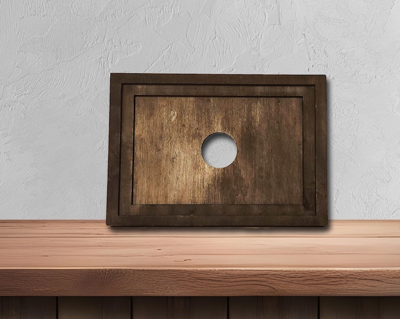 May include: A rectangular wooden board with a dark brown frame and a lighter brown center. A circular hole is cut in the center of the board. The board is displayed on a wooden surface.