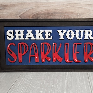 May include: A framed wooden sign with the text "Shake Your Sparkler" in white and red letters on a blue background.