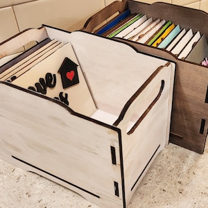 May include: Two rectangular wooden storage crates with cutout handles. The front crate is finished in a light whitewash, while the rear crate features a dark wood stain. Both contain stacked rectangular wooden signs with visible text and graphics.