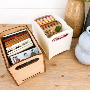 May include: Two wooden storage crates filled with assorted rectangular cards. One crate is natural light wood, the other is painted white. The cards feature various designs, including a green clover and the text "YOU ARE MY".