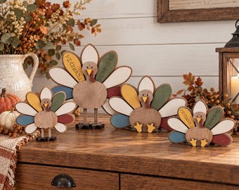 May include: A set of four decorative wooden turkey figures in varying sizes, featuring brown bodies and colorful tail feathers in shades of cream, blue, yellow, green, and red. Two of the turkeys have the words THANKFUL and GRATEFUL printed on feathers.