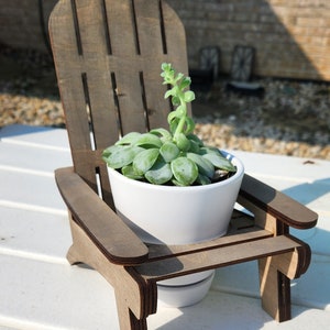 May include: A small wooden adirondack chair with a white planter holding a succulent plant. The chair and planter are sitting on a white surface.
