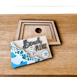 May include: Wooden beach-themed sign with the words "Beach PLEASE" in gray script. The sign features a wave design with blue accents, a starfish, and a sand dollar. A wooden frame with a hole is behind the sign.