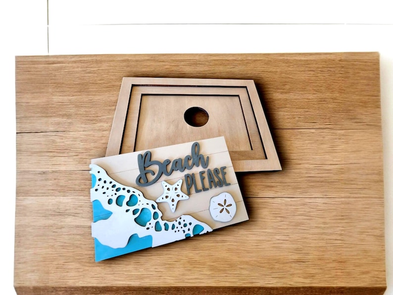 May include: Wooden beach-themed sign with the words "Beach PLEASE" in gray script. The sign features a wave design with blue accents, a starfish, and a sand dollar. A wooden frame with a hole is behind the sign.