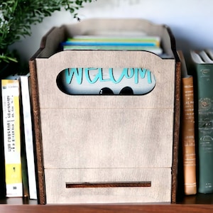 May include: A wooden box with a cutout that says "Welcome" in teal lettering. The box is filled with books and is sitting on a shelf with other books.