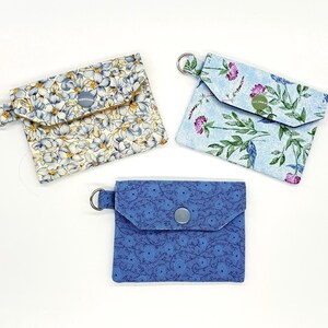 May include: Three small, square fabric pouches with snap closures and key rings. The pouches feature floral patterns in blue, white, and purple. One pouch is solid blue with a circular pattern.