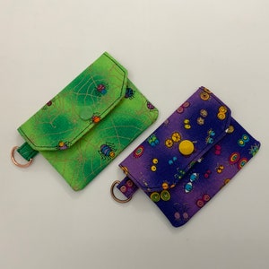 May include: Two small fabric coin purses with a zipper closure. One purse is green with a spider web pattern and the other is purple with a colorful eye pattern.