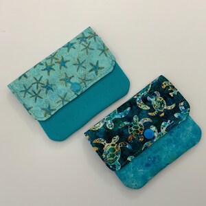 May include: Two rectangular fabric pouches with snap closures. One pouch features a light blue starfish pattern with a solid teal base. The second pouch displays a dark blue sea turtle pattern with a mottled blue and teal base.