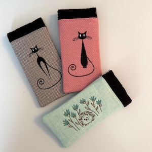 May include: Three rectangular fabric cases with black top edges. Two cases feature a white polka dot pattern with a black cat illustration, one in gray and one in pink. The third case is light green with a hedgehog and flower embroidery.