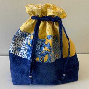 May include: A drawstring bag with a patchwork design. The bag features a navy blue base, a section with blue floral patterns on white, and yellow floral fabric at the top. A navy blue drawstring secures the top.