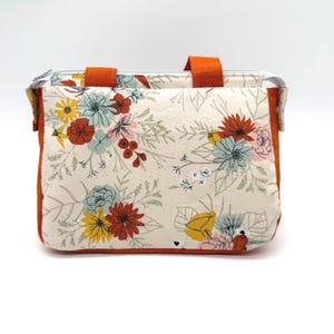 May include: A rectangular floral print bag with orange handles and trim. The bag features a cream-colored background with a colorful floral pattern in shades of red, yellow, blue, and pink. The bag has a zipper closure.
