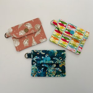May include: Three small fabric pouches with different designs. The first pouch is pink with a seashell pattern. The second pouch is white with a colorful fish pattern. The third pouch is blue with a sea turtle pattern. Each pouch has a small button closure.