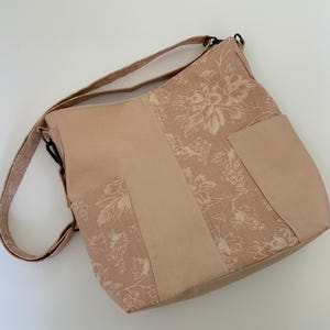 May include: A light pink shoulder bag with a floral pattern and two exterior pockets. The bag features a long adjustable strap and a solid light pink center panel. The floral design includes grape clusters and leaves.