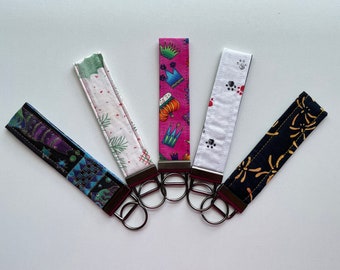 May include: Five colorful keychains with different patterns. The keychains are made of fabric and have metal rings. The patterns include a crown, pumpkins, paw prints, a floral design, and a geometric design.