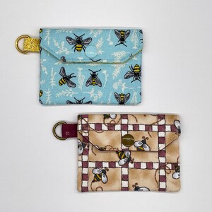 May include: Two small fabric pouches with bee designs. The pouch on the left is blue with a yellow button closure and a bee pattern. The pouch on the right is brown with a red button closure and a checkered pattern with bees.