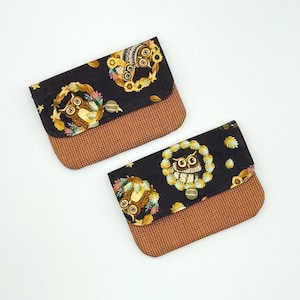 May include: Two small, rectangular wallets with a flap closure. The wallets feature a black background with an owl design surrounded by a floral wreath. The bottom half of the wallets is a brown, textured fabric. Each wallet has a metal snap closure.