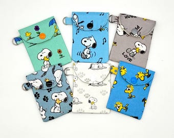 May include: Six small fabric pouches featuring Snoopy and Woodstock characters. Each pouch has a flap with a snap button closure and a metal D-ring on the side. The designs include light green, blue, gray, and white patterns with various cartoon prints.