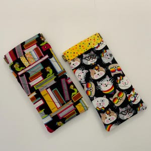 May include: Two rectangular fabric cases with metal frames. The left case features a black background with a colorful stack of books and green worms. The right case has a black background with cat faces wearing rainbow glasses and a yellow polka dot top.