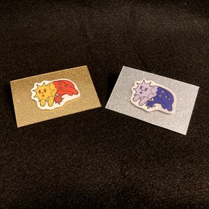 Op de afbeelding: Twee emaille-pins op displaykaarten. De pin aan de linkerkant is een gele en rode kat met een zonontwerp. De pin aan de rechterkant is een paarse kat met een maan- en sterrendesign.