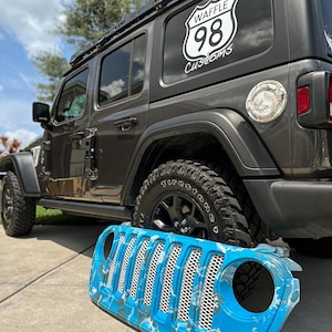 May include: A gray Jeep Wrangler with a black roof rack and black wheels. A custom blue and white front grill is in front of the vehicle. The rear window has a white logo that says "WAFFLE 98 Customs".
