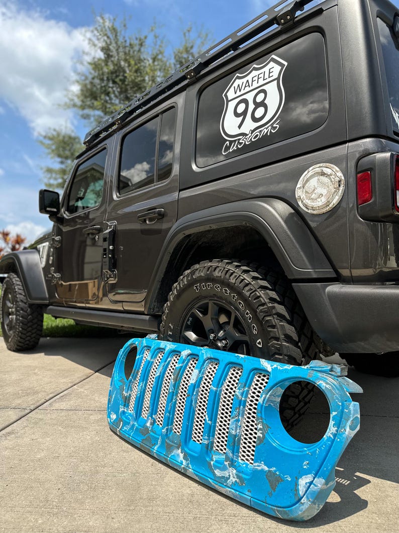 May include: A gray Jeep Wrangler with a black roof rack and black wheels. A custom blue and white front grill is in front of the vehicle. The rear window has a white logo that says "WAFFLE 98 Customs".
