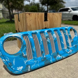 May include: A custom blue and white marbled Jeep grille. The grille has a honeycomb mesh design and is set against a wooden crate and a suburban backdrop.