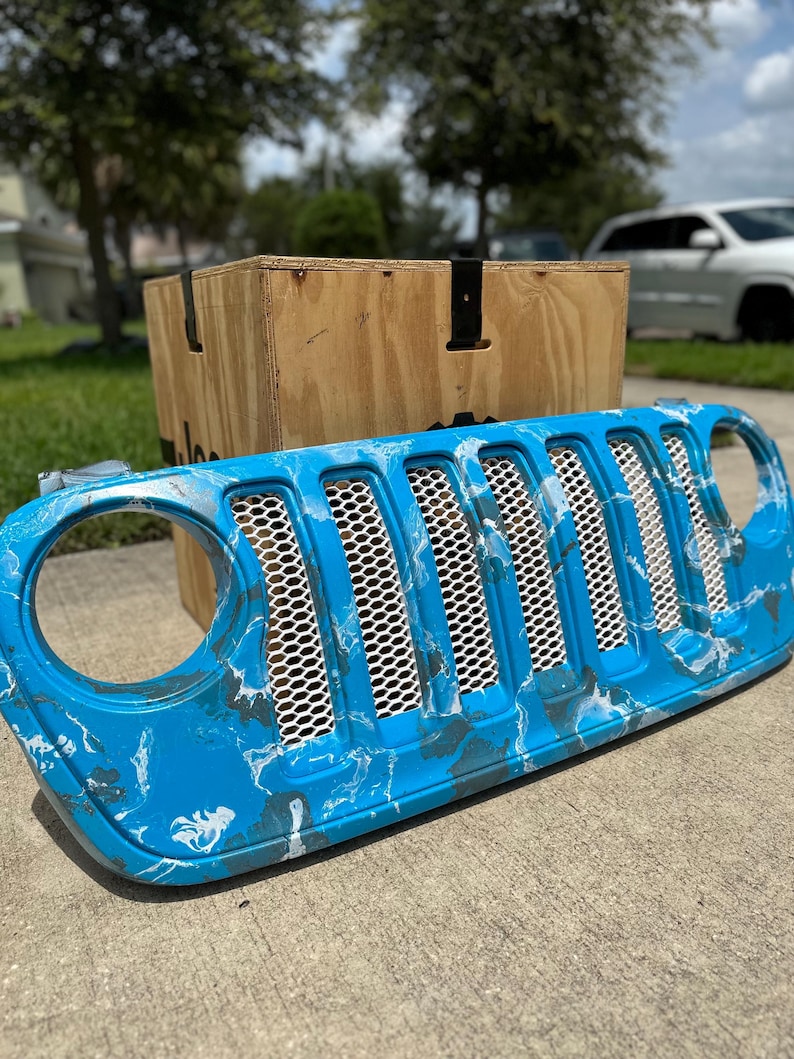 May include: A custom blue and white marbled Jeep grille. The grille has a honeycomb mesh design and is set against a wooden crate and a suburban backdrop.
