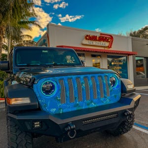 May include: Black Jeep Wrangler with a custom blue and white grill design. The vehicle has large off-road tires, a black bumper, and LED headlights. The background includes a blue sky and a building with a sign that says "Chill Ice Cream".
