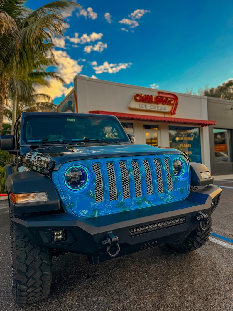 May include: Black Jeep Wrangler with a custom blue and white grill design. The vehicle has large off-road tires, a black bumper, and LED headlights. The background includes a blue sky and a building with a sign that says "Chill Ice Cream".