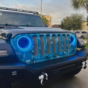 May include: A modified blue Jeep with a custom front grille and blue halo headlights. The grille has a blue and white marbled design. The front bumper has white tow hooks and additional lights. A roof rack is also visible.