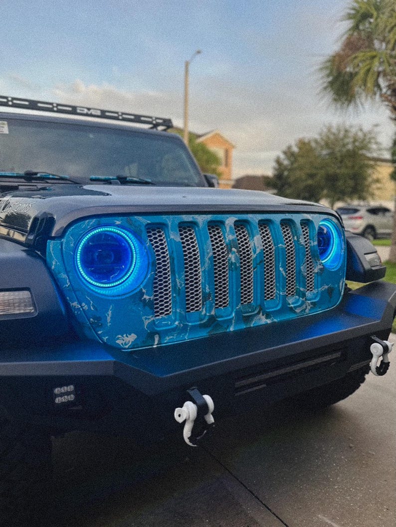 May include: A modified blue Jeep with a custom front grille and blue halo headlights. The grille has a blue and white marbled design. The front bumper has white tow hooks and additional lights. A roof rack is also visible.