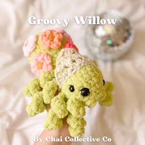 May include: A green crocheted spider with a white crocheted hat and pink and orange flowers on its head. The spider has black eyes and a friendly expression. The text "Groovy Willow" is written above the spider and "By Chai Collective Co" is written below the spider.