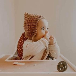 May include: A baby wearing a brown knitted bonnet and a white long-sleeved shirt sits in a high chair. The high chair has a red and white polka dot seat cover. A pacifier and a snack are on the tray.