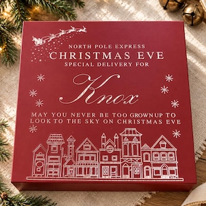 Könnte beinhalten: Eine quadratische rote Geschenkbox mit weißem Text, der North Pole Express Christmas Eve Special Delivery For Knox lautet, sowie dem Satz May you never be too grownup to look to the sky on Christmas Eve und einer Strichzeichnung eines Dorfes.