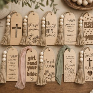 May include: A collection of ten light-colored wooden bookmarks with rounded tops, each featuring unique engraved text, floral illustrations, or cross cutouts. Most include natural jute tassels with wooden beads, while two feature fabric ribbons.