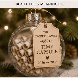 May include: A clear glass spherical ornament filled with numerous small, rolled-up paper scrolls. A rectangular wooden tag hangs from the top, engraved with the text: THE TACKETT FAMILY TIME CAPSULE 2026 → 2036, featuring a small heart and leaf icon.