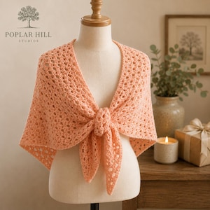 May include: A peach-colored triangular shawl featuring an intricate openwork crochet pattern. The garment is draped over a mannequin and tied in a knot at the center front, showcasing the delicate texture and consistent yarn color throughout.