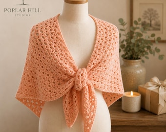 May include: A peach-colored triangular shawl featuring an intricate openwork crochet pattern. The garment is draped over a mannequin and tied in a knot at the center front, showcasing the delicate texture and consistent yarn color throughout.