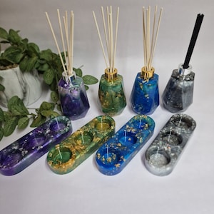 Könnte beinhalten: Vier geometrische Glasdiffusor-Flaschen mit Holz- und schwarzen Schilfrohren und drei passende ovale Diffusor-Ständer mit drei Vertiefungen. Die Flaschen sind mit farbigem Harz und goldenen Flocken gefüllt. Die Ständer sind lila, grün, blau und grau.