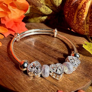 Könnte beinhalten: Ein silbernes Armband mit einem Doppelstrang-Band und vier Anhängern. Die Anhänger sind herzförmig mit filigranen Designs und einer silbernen Perle.