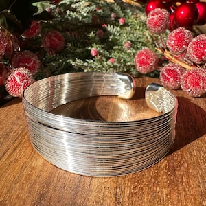 May include: A silver bangle bracelet made of multiple thin wires woven together. The bracelet is on a wooden surface.