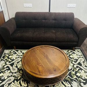 May include: A round wooden coffee table with intricate carvings sits on a black and white patterned rug in front of a brown sofa.