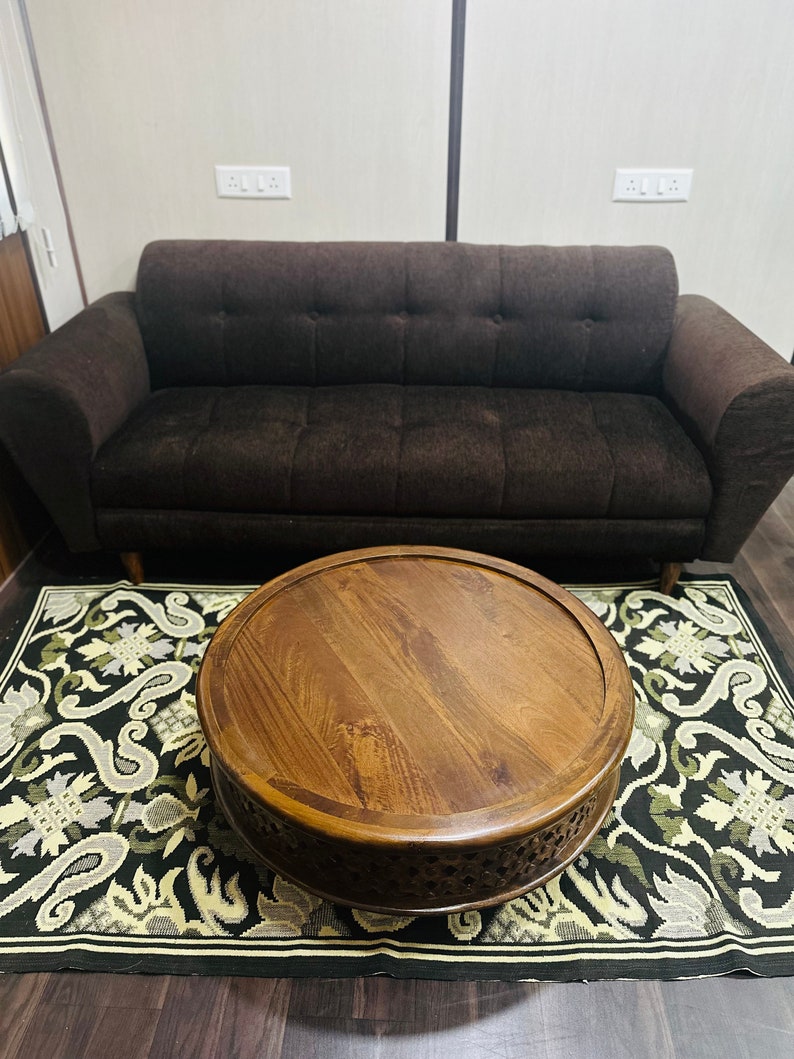 May include: A round wooden coffee table with intricate carvings sits on a black and white patterned rug in front of a brown sofa.