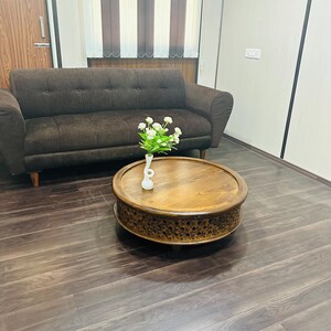 May include: A brown leather sofa with a wooden coffee table in front of it. The coffee table has a carved design and a white vase with white flowers on top. The room has a wooden floor and white walls.
