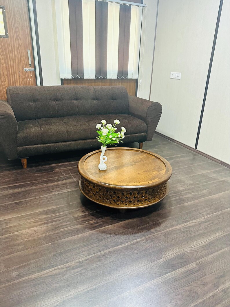 May include: A brown leather sofa with a wooden coffee table in front of it. The coffee table has a carved design and a white vase with white flowers on top. The room has a wooden floor and white walls.
