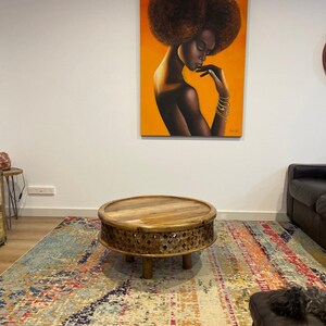 May include: A round wooden coffee table with intricate carvings around the edge and three legs. The table is on a colorful rug with a painting of a woman with an afro hairstyle on the wall behind it.