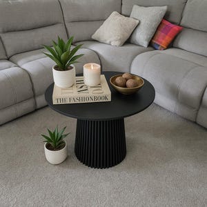 Puede incluir: Una mesa de centro redonda negra con base de listones, que sostiene una vela encendida, un cuenco decorativo con tres esferas y un libro titulado "THE FASHIONBOOK". Dos pequeñas plantas en maceta están cerca, con un sofá seccional gris en el fondo.