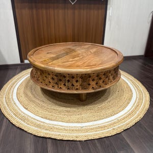 Pode incluir: Uma mesa de centro redonda de madeira com entalhes intrincados ao redor da borda inferior. A mesa está sobre um tapete de fibra natural com listras brancas.