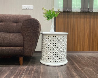 Puede incluir: Mesa auxiliar de madera blanca con un diseño decorativo de corte. La mesa tiene una tapa redonda y una base cilíndrica. Un jarrón con flores blancas se coloca encima de la mesa.