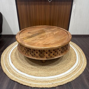 May include: A round wooden coffee table with intricate carvings around the base. The table is sitting on a natural fiber rug with white trim.